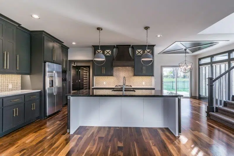 Justina II Floor Plan Kitchen With Large Island, Dark Cabinetry, Hardwood Floors, And Modern Pendant Lighting By Comerio Homes