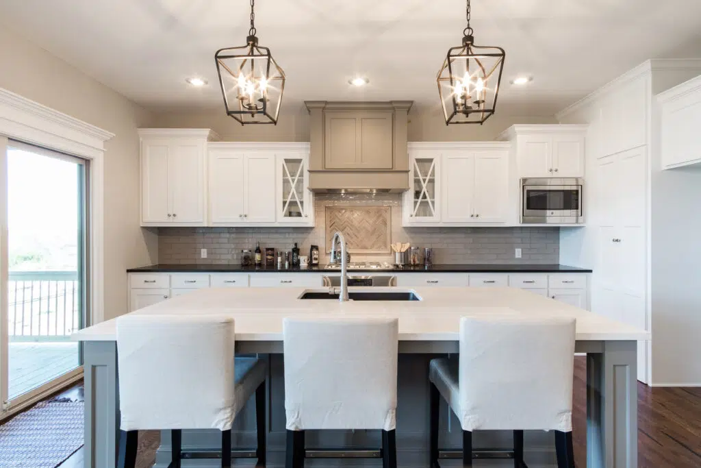 Comerio Homes Kitchen Featuring A Large Island, White Cabinetry, Pendant Lighting And A Decorative Range Hood.