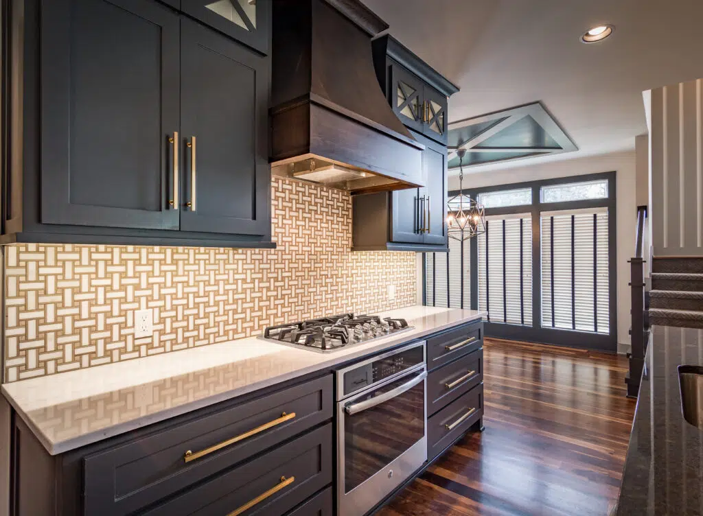 Justina Floor Plan Kitchen By Comerio Homes Featuring A Custom Wood Range Hood, Dark Cabinetry, Brass Hardware And Patterned Backsplash.