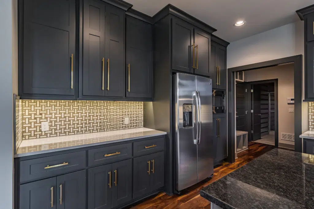 Justina Floor Plan Kitchen By Comerio Homes With Dark Cabinetry, Brass Hardware, Patterned Backsplash And Stainless Steel Refrigerator.