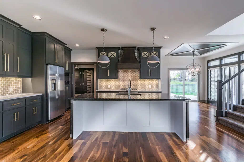 Justina Floor Plan Kitchen By Comerio Homes Featuring A Large Island, Dark Cabinetry, Patterned Backsplash And Pendant Lighting.