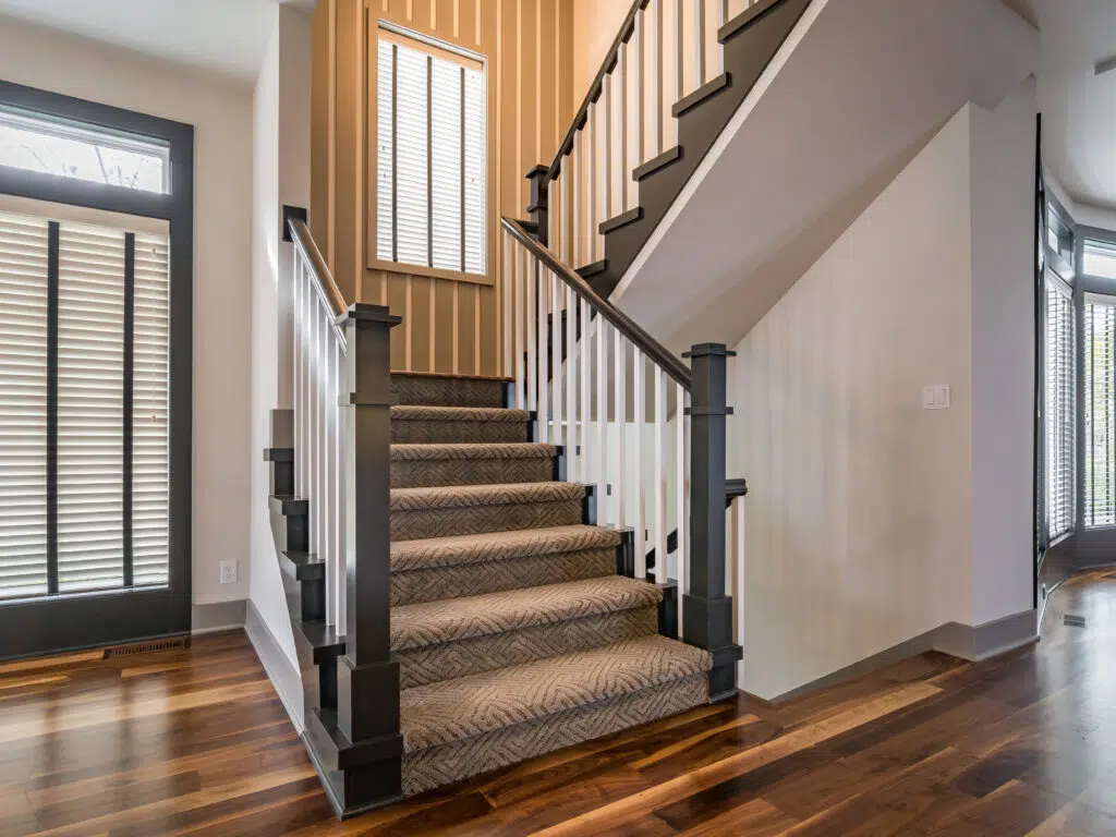 Justina Floor Plan Staircase By Comerio Homes With Carpeted Stairs, Dark Wood Railings, White Balusters And Hardwood Flooring.